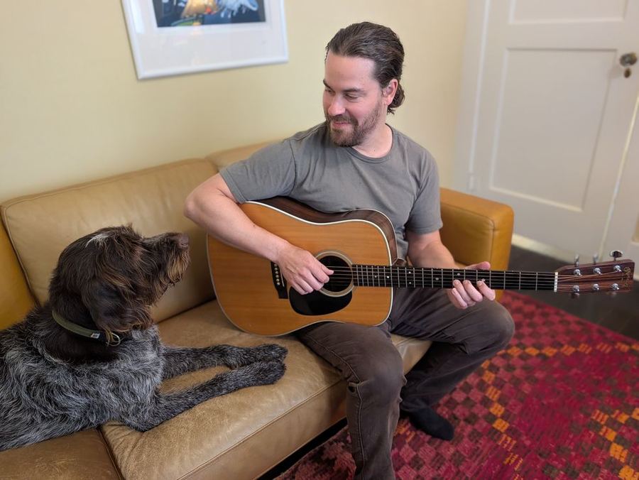 David Zuckerman playing acoustic guitar at home in Santa Rosa, with his dog listening nearby